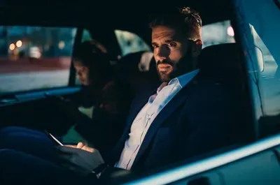 Man in suit, looking concerned, using phone in car. Dark, nighttime setting.