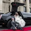 Bride in white dress sits in a black limo with groom looking up at her, on a red carpet.