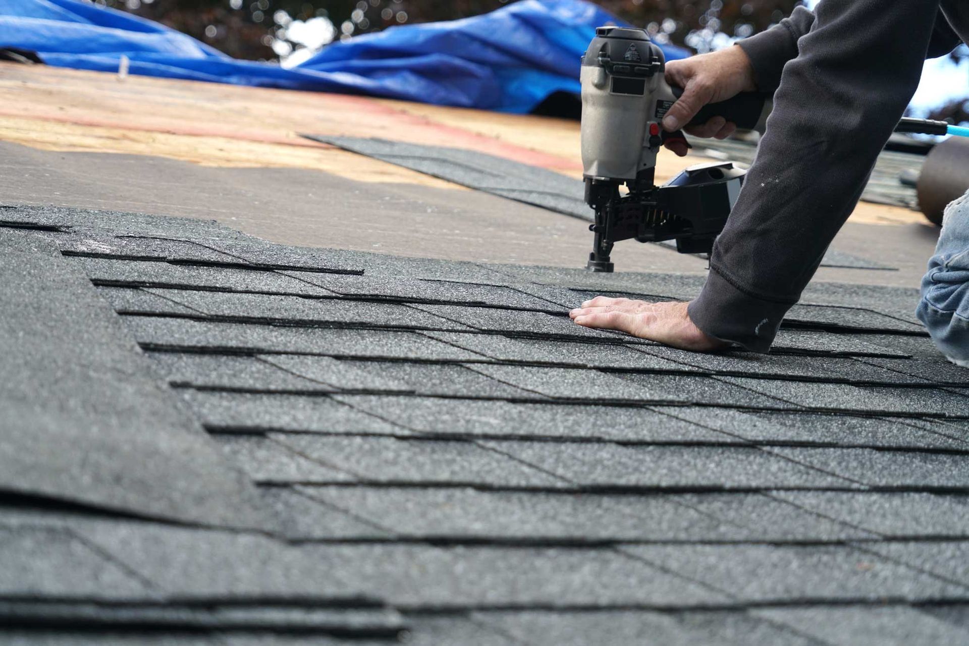 Roofer installing asphalt shingles with a nail gun on a roof surface.