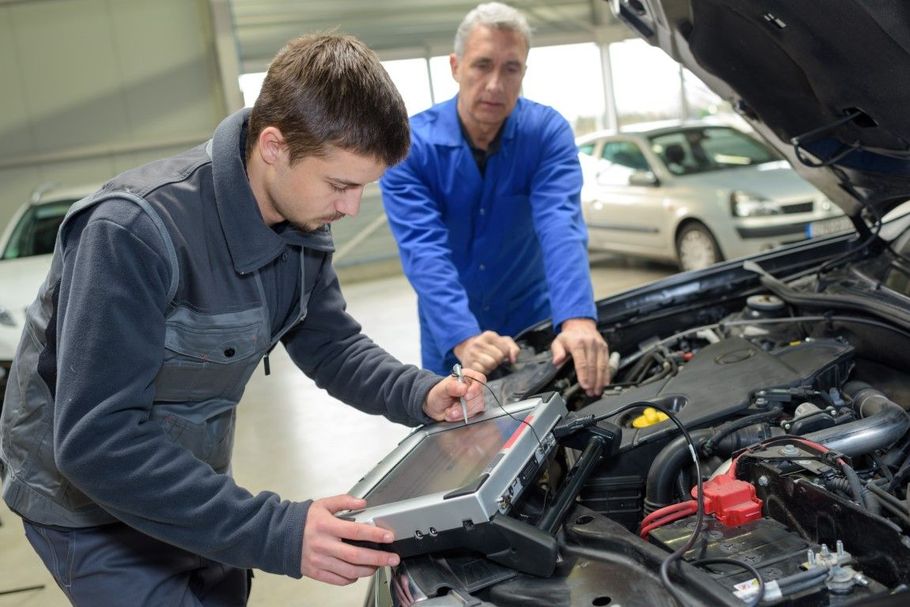 Two mechanics inspect an open car engine while one holds a diagnostic laptop.