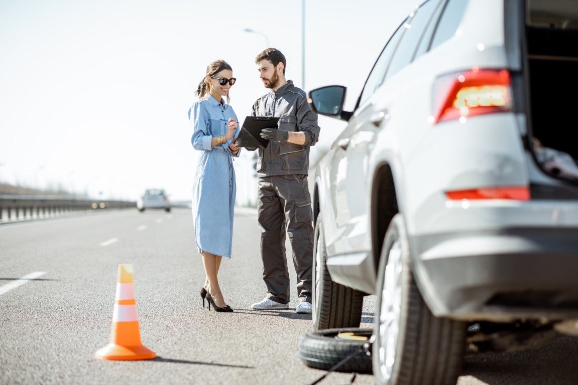 Driver and roadside assistant beside a broken-down silver SUV on the highway, with an orange warning cone.