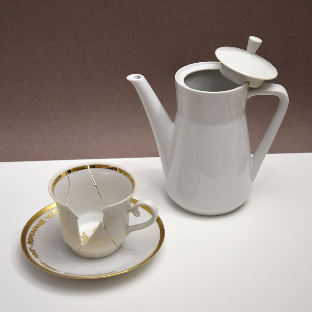 Titel: Zu spät
Ansicht seitlich.
Die Plastik der Künstlerin Benita von Wendt besteht
aus einer Tasse der ein großes Stück Porzellan fehlt. In der Tasse steht der Schriftzug Hass mit Blattgold geschrieben. Daneben steht ein Kännchen, das von Innen beleuchtet ist. Innen befindet sich der Schriftzug Toleranz.