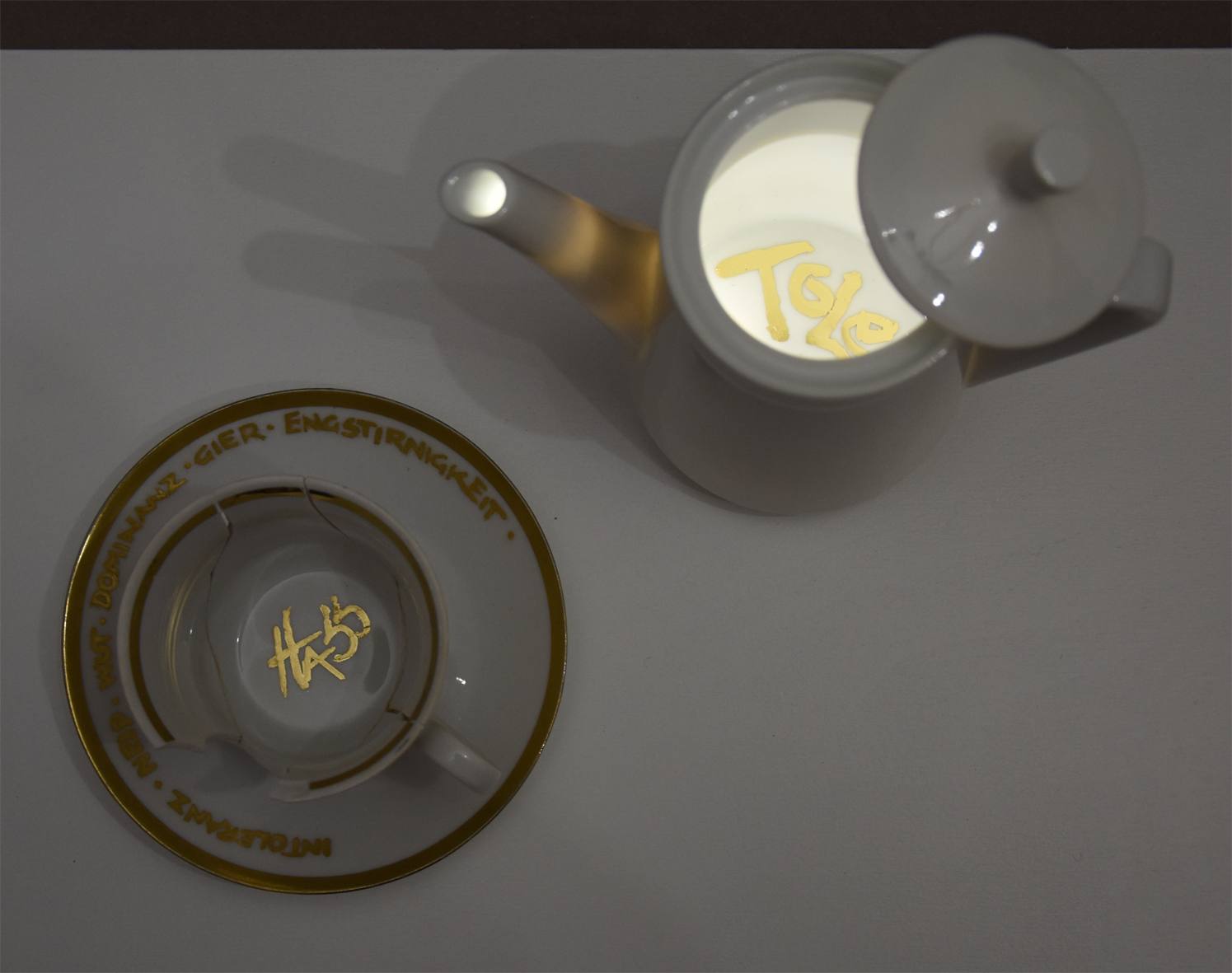 Titel: Zu spät
Ansicht Ensemble von oben beleuchtet.
Die Plastik der Künstlerin Benita von Wendt besteht
aus einer Tasse der ein großes Stück Porzellan fehlt. In der Tasse steht der Schriftzug Hass mit Blattgold geschrieben. Daneben steht ein Kännchen, das von Innen beleuchtet ist. Innen befindet sich der Schriftzug Toleranz.