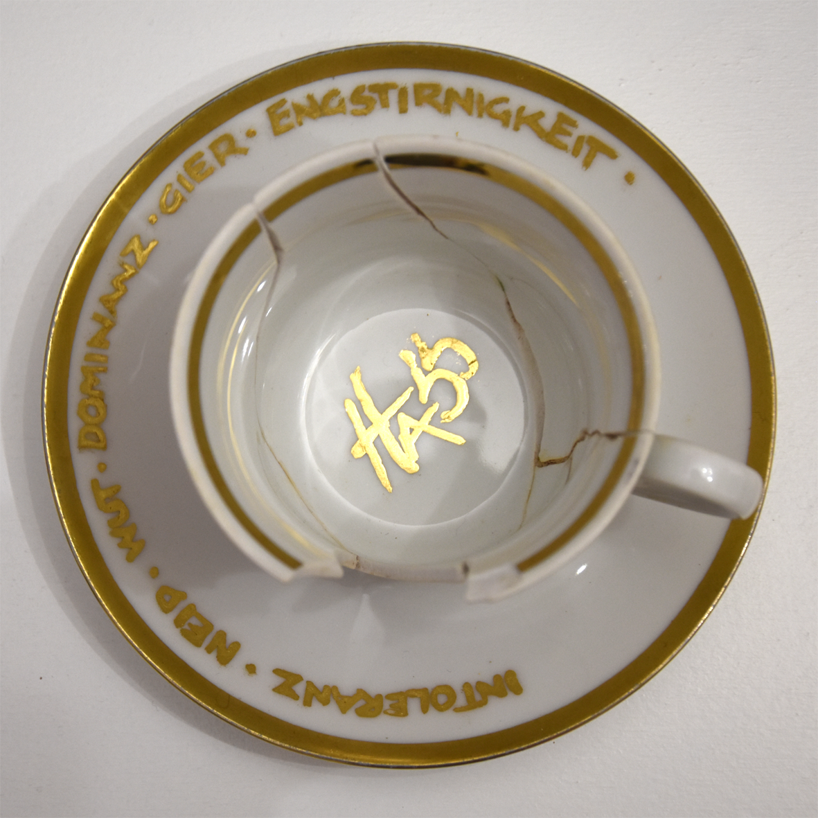 Titel: Zu spät
Ansicht Detail Tasse von oben.
Die Plastik der Künstlerin Benita von Wendt besteht
aus einer Tasse der ein großes Stück Porzellan fehlt. In der Tasse steht der Schriftzug Hass mit Blattgold geschrieben. Daneben steht ein Kännchen, das von Innen beleuchtet ist. Innen befindet sich der Schriftzug Toleranz.