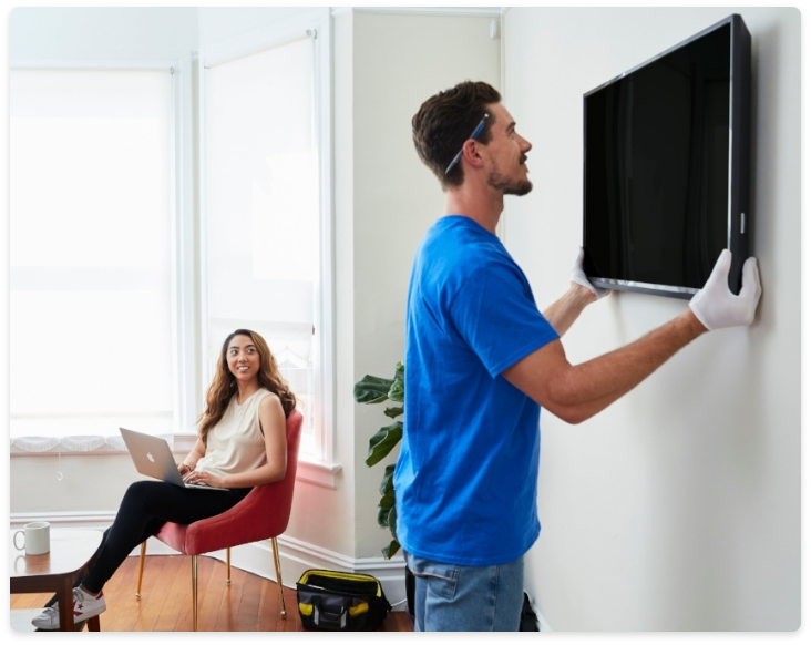 A man is installing a flat screen tv on a wall while a woman sits in a chair with a laptop.