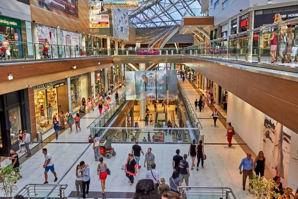 Busy indoor shopping mall with people walking beneath a glass roof and storefronts on both sides