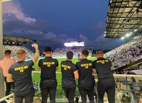 Five staff in black “SCOTT’S” shirts watch a lit stadium crowd at dusk.