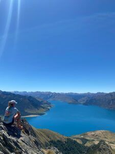 Person sitting on a rocky cliff overlooking a bright blue lake and distant mountains under a clear sky