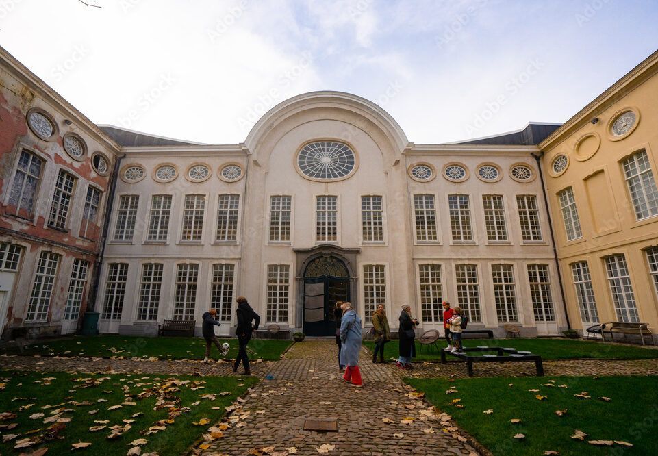 Grand historic building courtyard with tall windows, central archway, and a few people walking on a leaf-strewn path