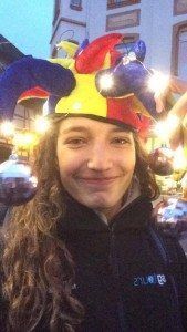 Person smiling in a red-and-yellow jester hat with silver bells indoors.