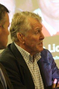 Elderly man in a dark jacket speaking in profile at a presentation, with a projected screen in the background