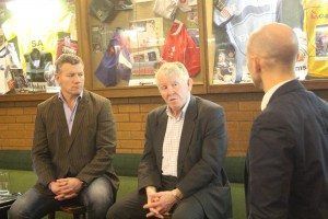 Three men seated and talking in a cozy indoor setting with framed photos on the wall.