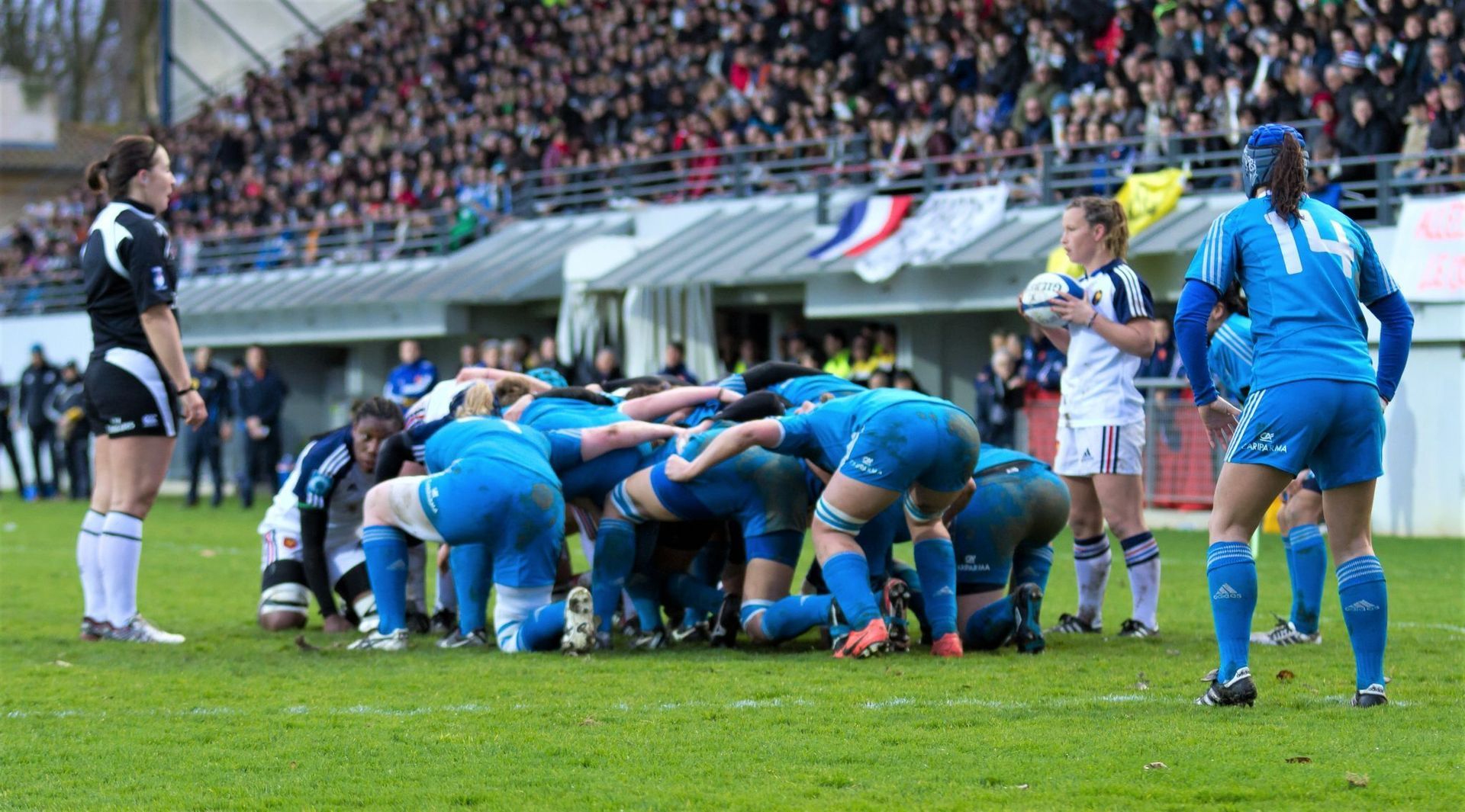 Women’s rugby scrum in blue uniforms, with crowd watching from the stands