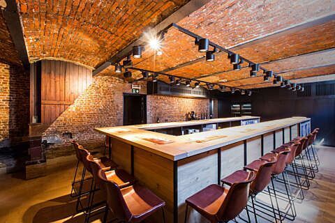 Brick-walled bar interior with a long wooden counter and red stools under warm pendant lights