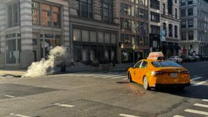 Orange taxi turning on a city street beside a plume of steam from a sidewalk vent