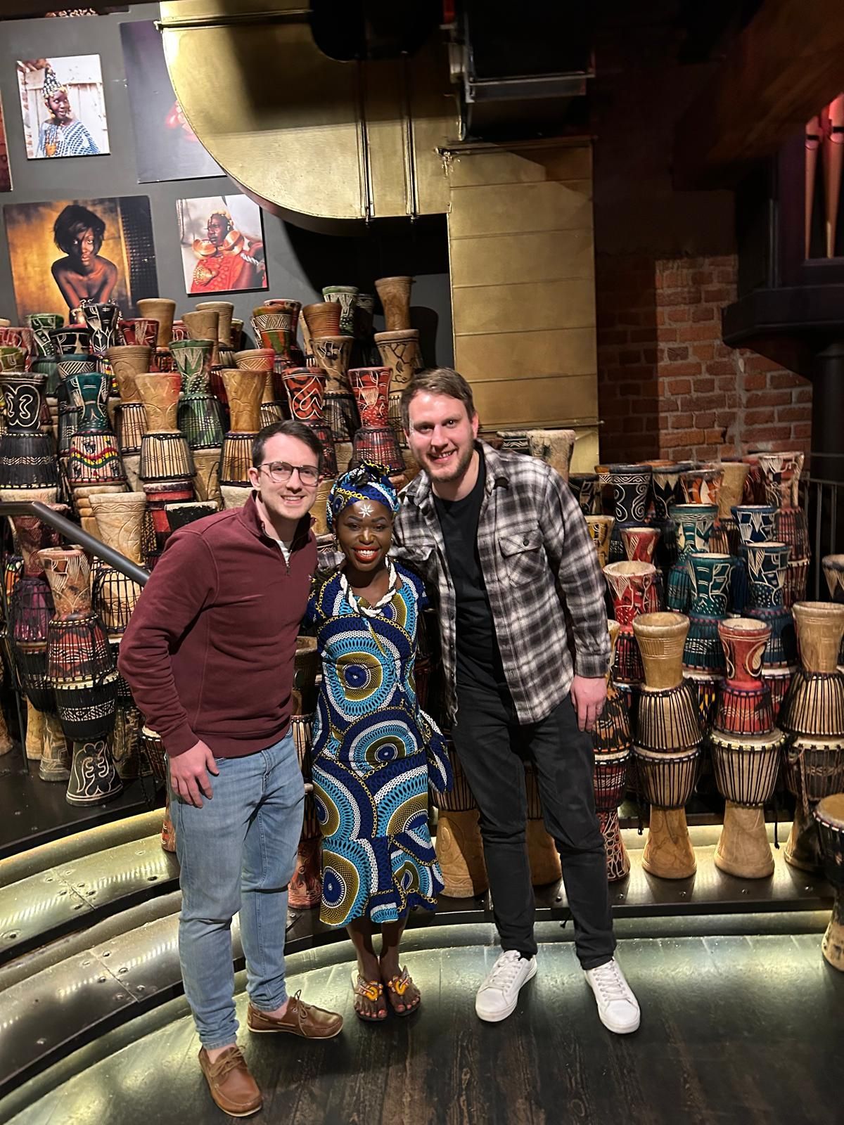 Three people pose in front of stacked drums in a colorful indoor shop or exhibit.