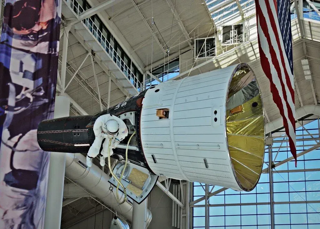 NASA spacecraft display with a white module, yellow interior, and American flag in a museum atrium