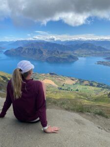 Person seated on a hillside overlooking a blue lake, mountains, and cloudy sky
