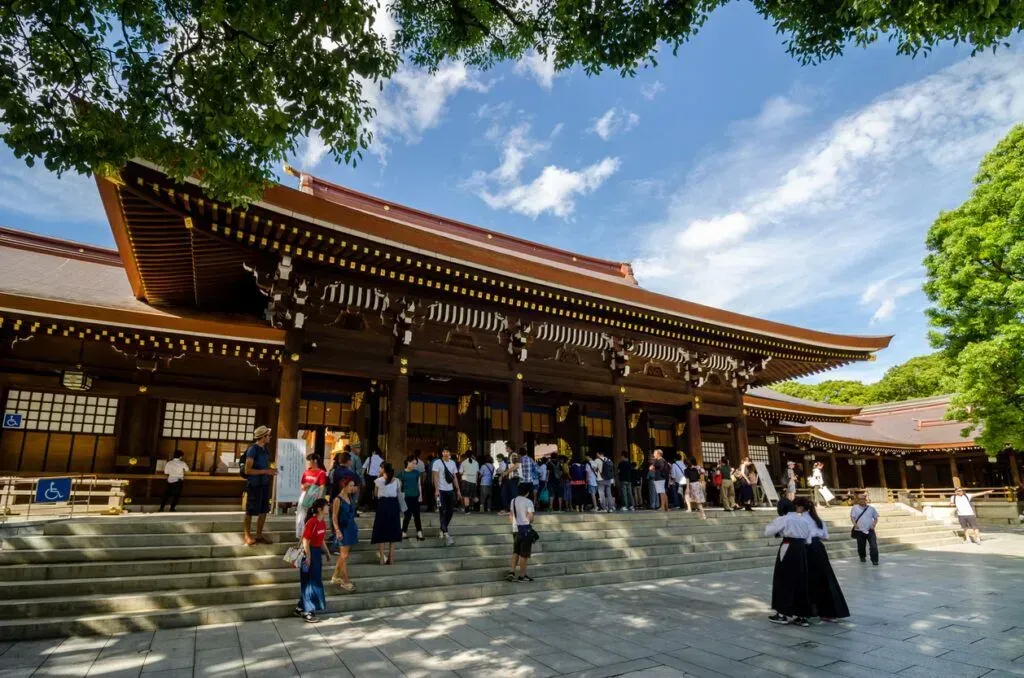 Temple courtyard with a large wooden gate, people gathered on the steps, and a blue sky overhead