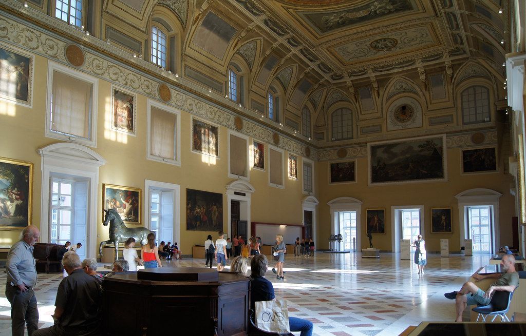 Grand ornate hall with marble floor, chandeliers, and a few people seated and standing around tables