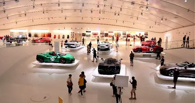 Car showroom with several sports cars displayed on a glossy floor under bright lights, with visitors walking around