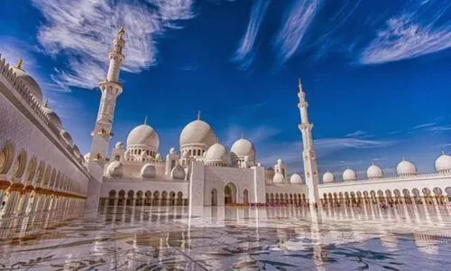 Grand white mosque with domes and minarets reflected on a polished courtyard under a blue sky