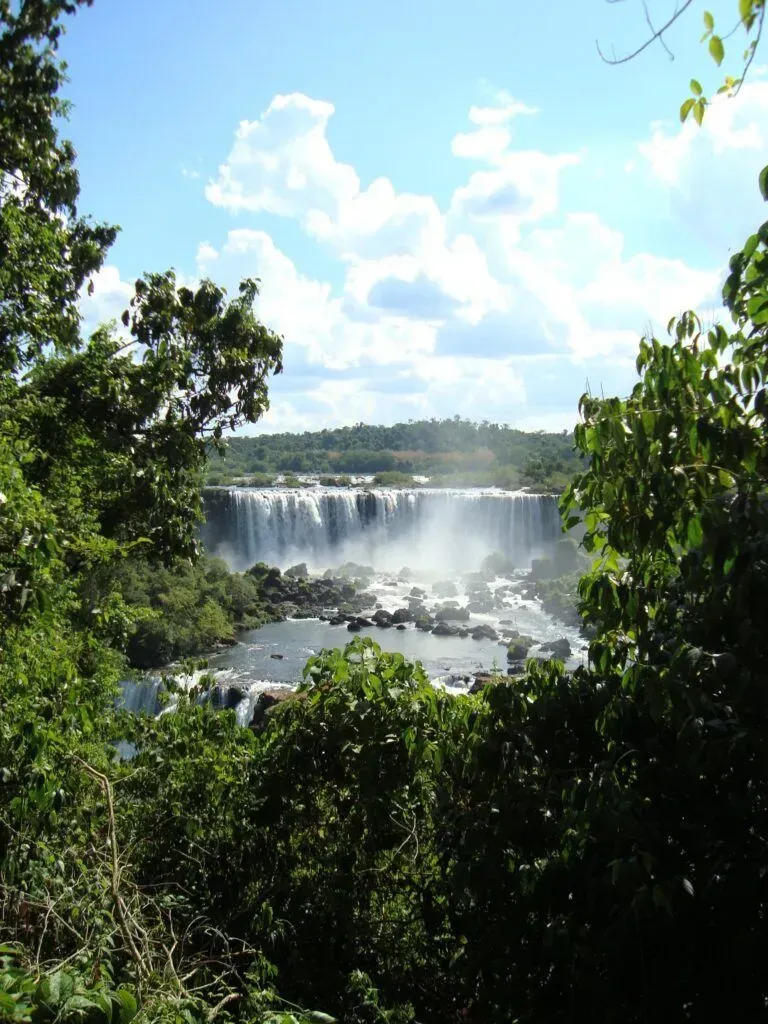 Wide waterfall cascading over rocks, framed by green trees under a bright blue sky.