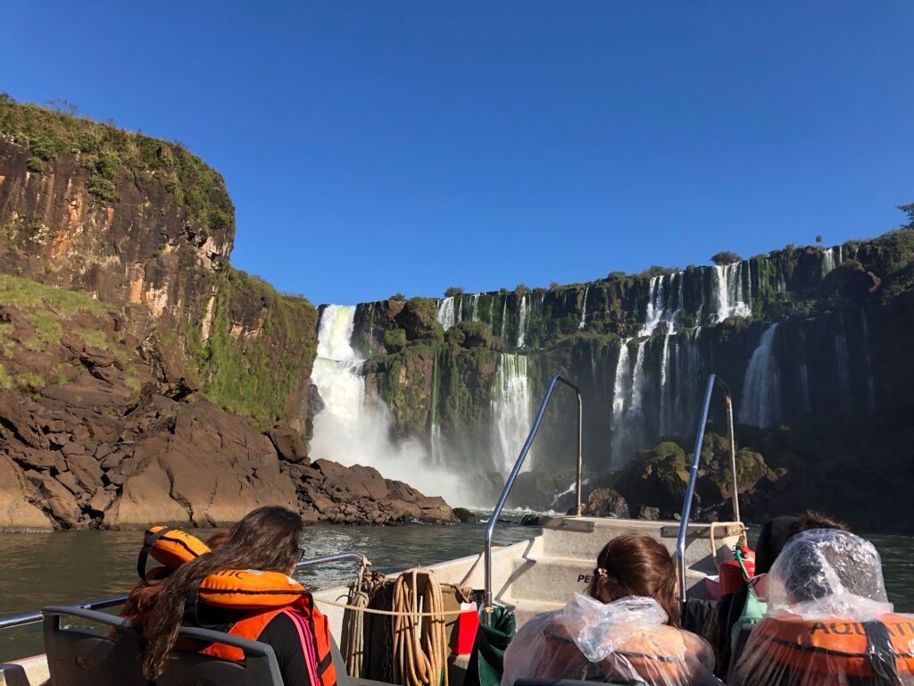 Tourists on a boat viewing a wide waterfall and rocky cliff under a clear blue sky