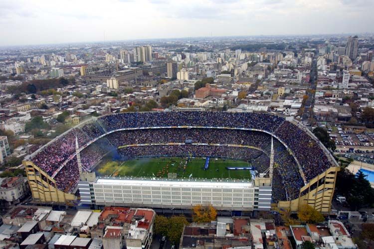 Aerial view of a packed soccer stadium in a city, with a green field and surrounding urban buildings.