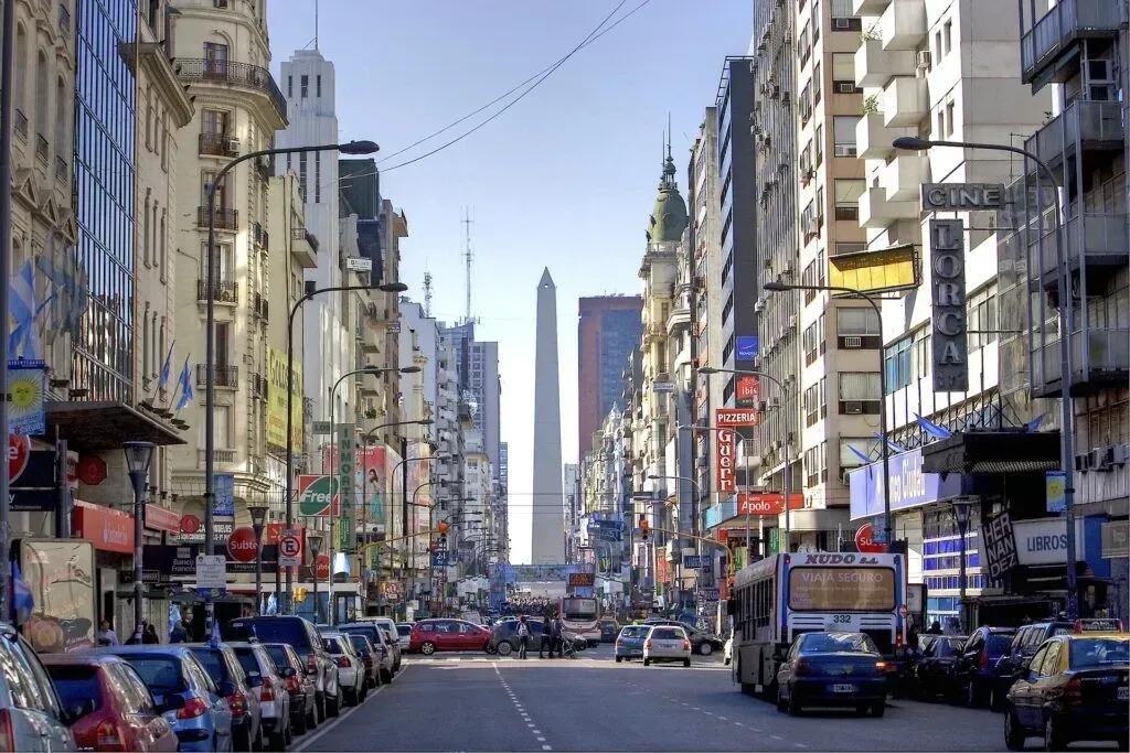 Busy city street lined with tall buildings, parked cars, and traffic under a clear sky
