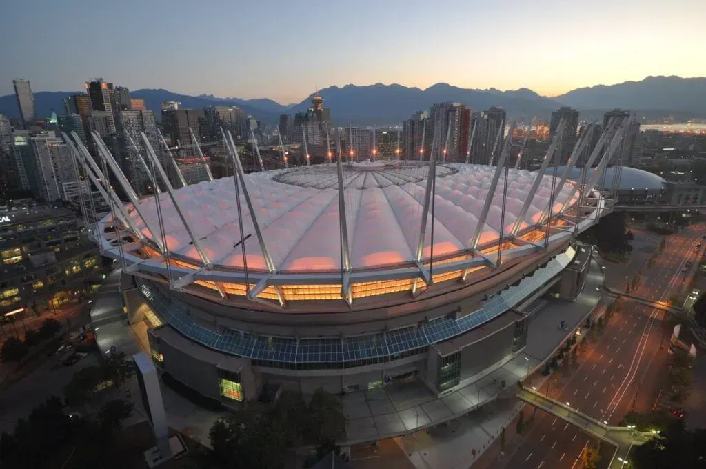 Aerial view of a stadium at dusk with glowing roof lights and city high-rises in the background