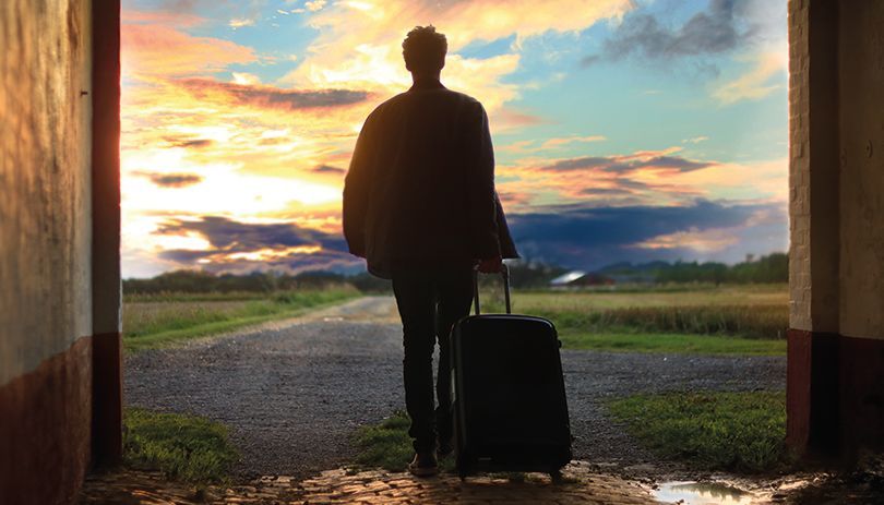 Silhouetted traveler with suitcase at a doorway, facing a colorful sunset over fields and clouds.
