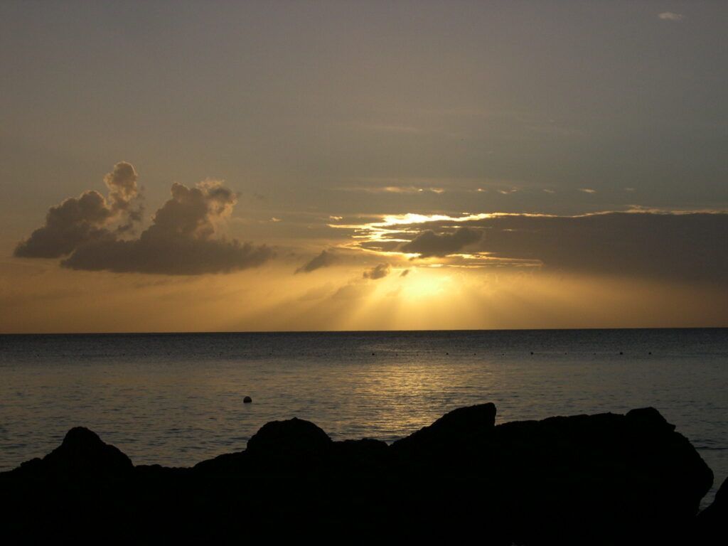 Sunset over calm ocean with golden light, dark clouds, and silhouetted rocks in the foreground