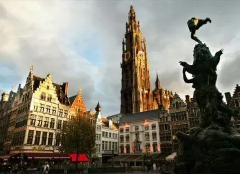Historic European square with ornate buildings and a tall cathedral tower beside a dark fountain statue