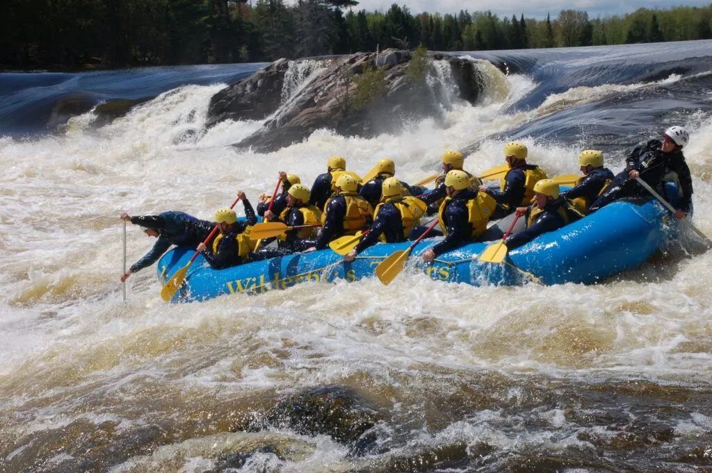 Group whitewater rafting in a blue raft through rough rapids beneath a waterfall