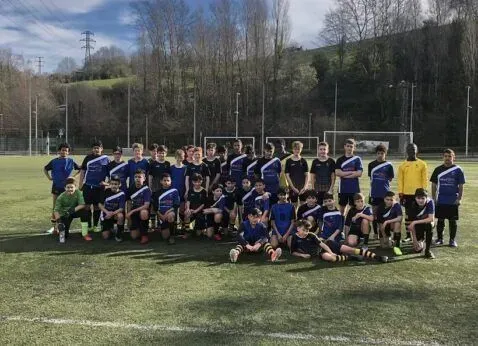 Soccer team posing on a grassy field, wearing blue and black uniforms with a goal and trees behind them.