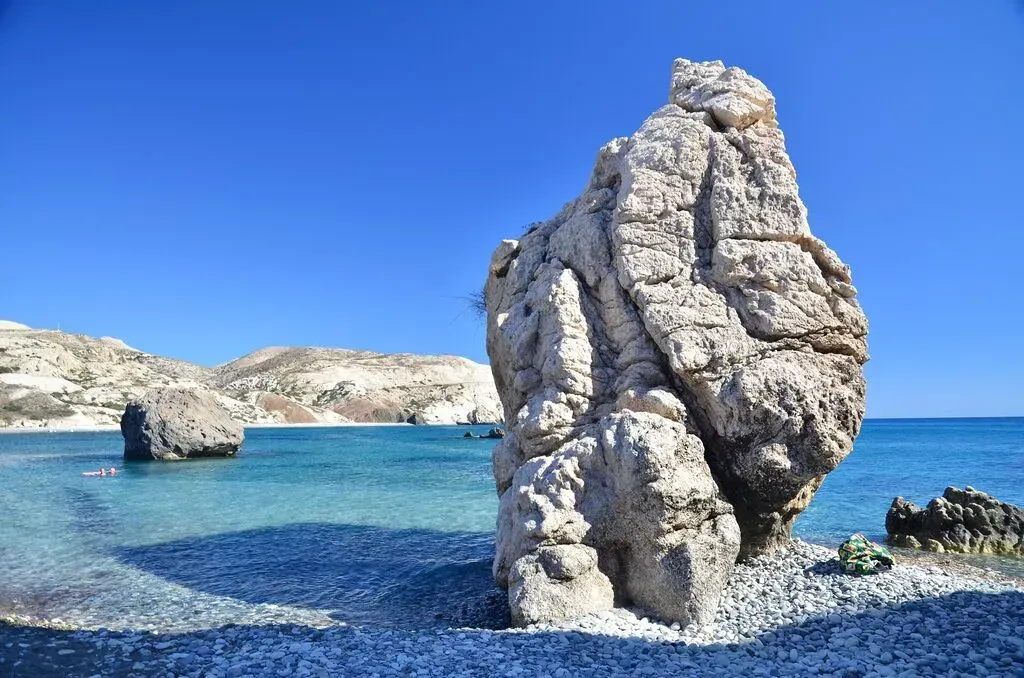 Large rock formation in bright blue shallow water along a rocky shoreline under clear skies