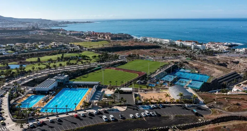 Aerial view of a coastal sports complex with blue tennis courts, parking lots, and the ocean in the background