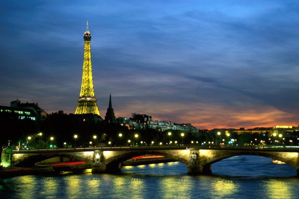 Eiffel Tower glowing at dusk over the Seine, with illuminated bridge and city lights reflected in the water