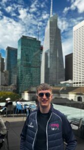 Man in sunglasses and dark jacket standing in front of tall city skyscrapers under a cloudy sky