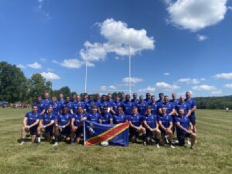 Large group in blue shirts posing on a field under a sunny sky, with a flag in front