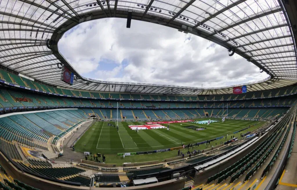 Wide-angle view of a covered stadium with green field and teal seating under a cloudy sky