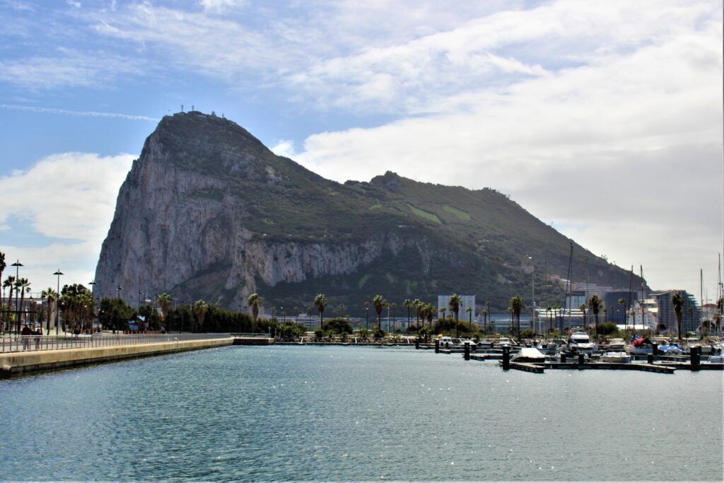 Coastal harbor with a steep rocky mountain, calm blue water, and boats under a cloudy sky
