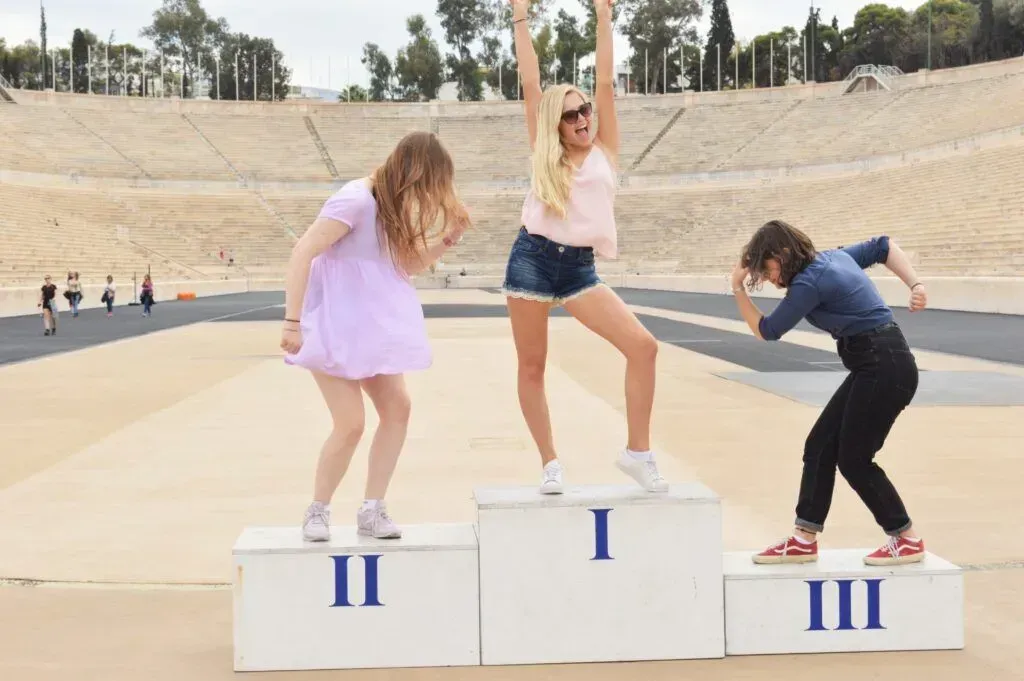 Three people on a winners' podium in a sandy outdoor bowl, posing playfully with raised arms.