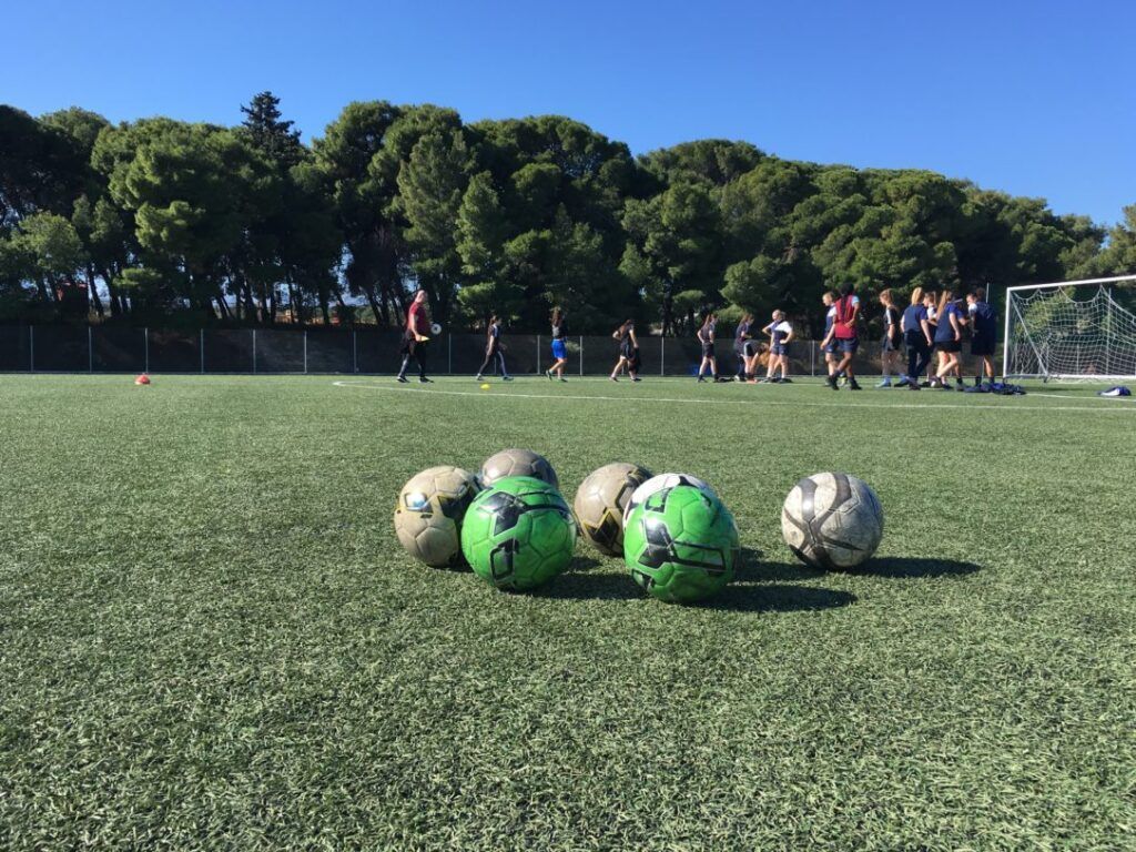 Soccer balls on turf with players gathered near a goal on a sunny field