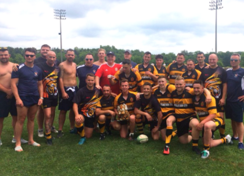 Rugby team posing on a grassy field, with players in black-and-gold jerseys and others shirtless, under cloudy skies