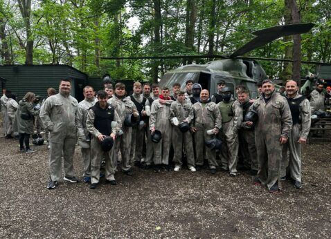 Group of people in camouflage gear posing outdoors beside a helicopter in a wooded area