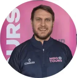 Man smiling in a navy MSQ Tours jacket against a pink branded backdrop