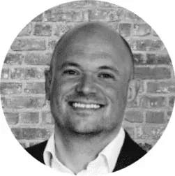 Black-and-white headshot of a smiling bald man in a suit before a brick wall