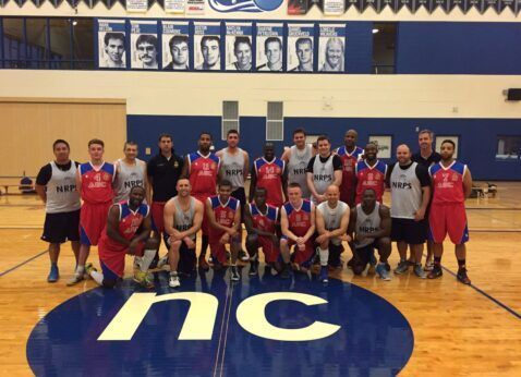 Group of basketball players and coaches posing on an indoor court with a large “NC” logo at center.